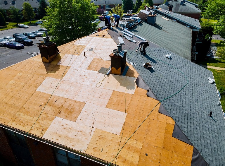 trzech mężczyzn wykonuje przebudowę dachu, układając na drewnianych płytach osb membranę dachową i papę bitumiczną