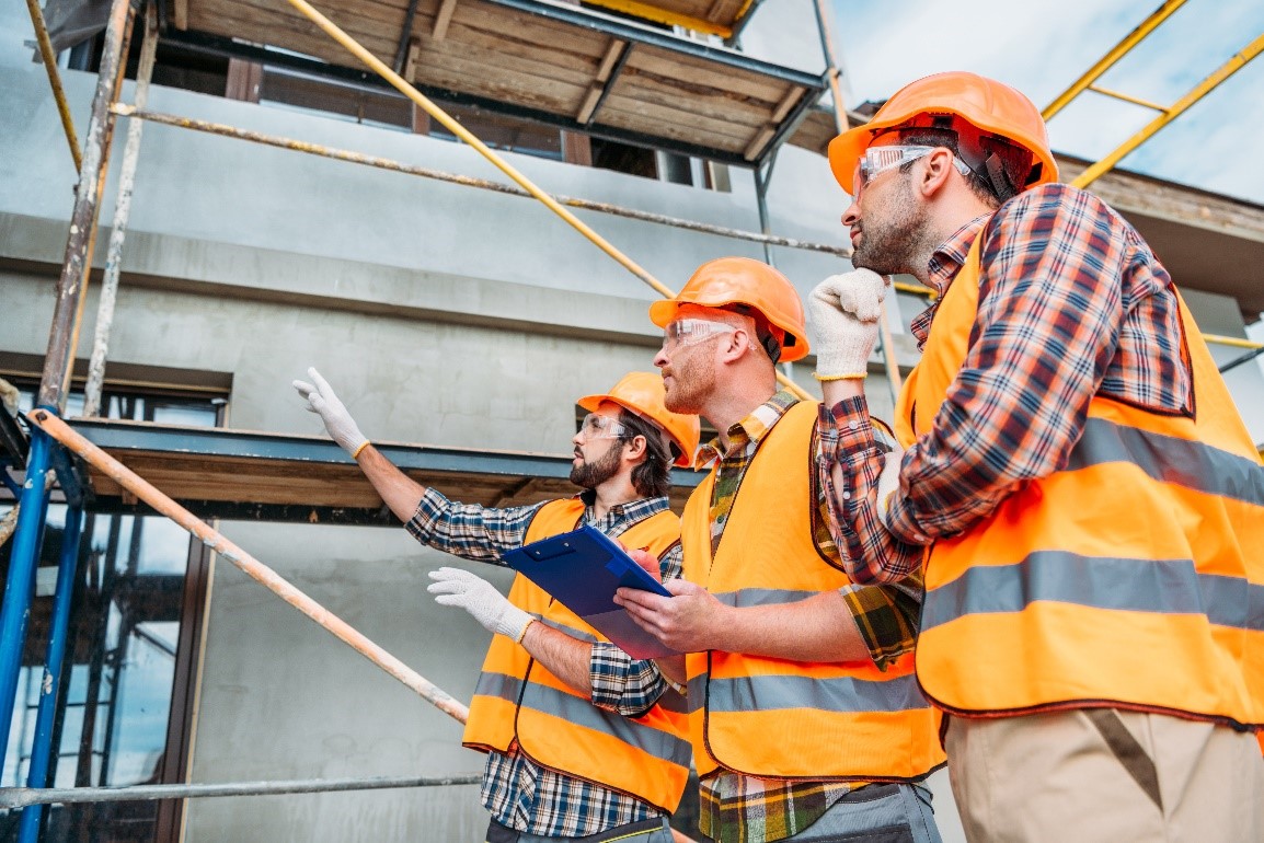 trzech młodych mężczyzn w pomarańczowych kaskach i kamizelkach odblaskowych, ocenia etap budowy domu