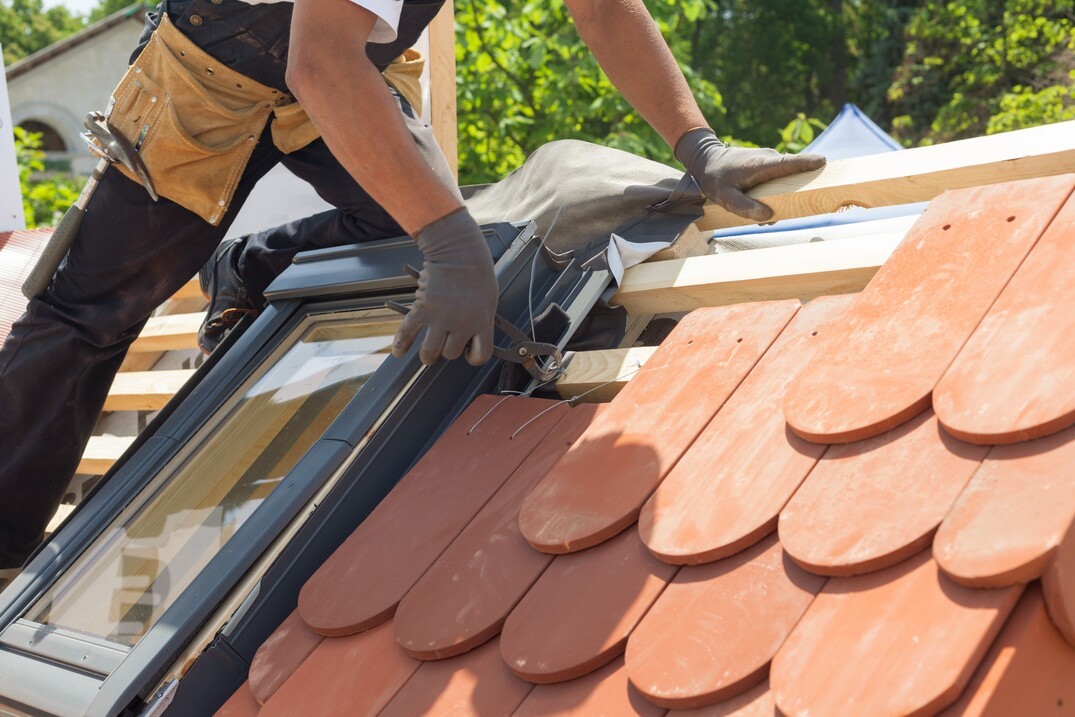 mężczyzna w kombinezonie roboczym układa na drewnianej konstrukcji dachu pomarańczową, betonową dachówkę