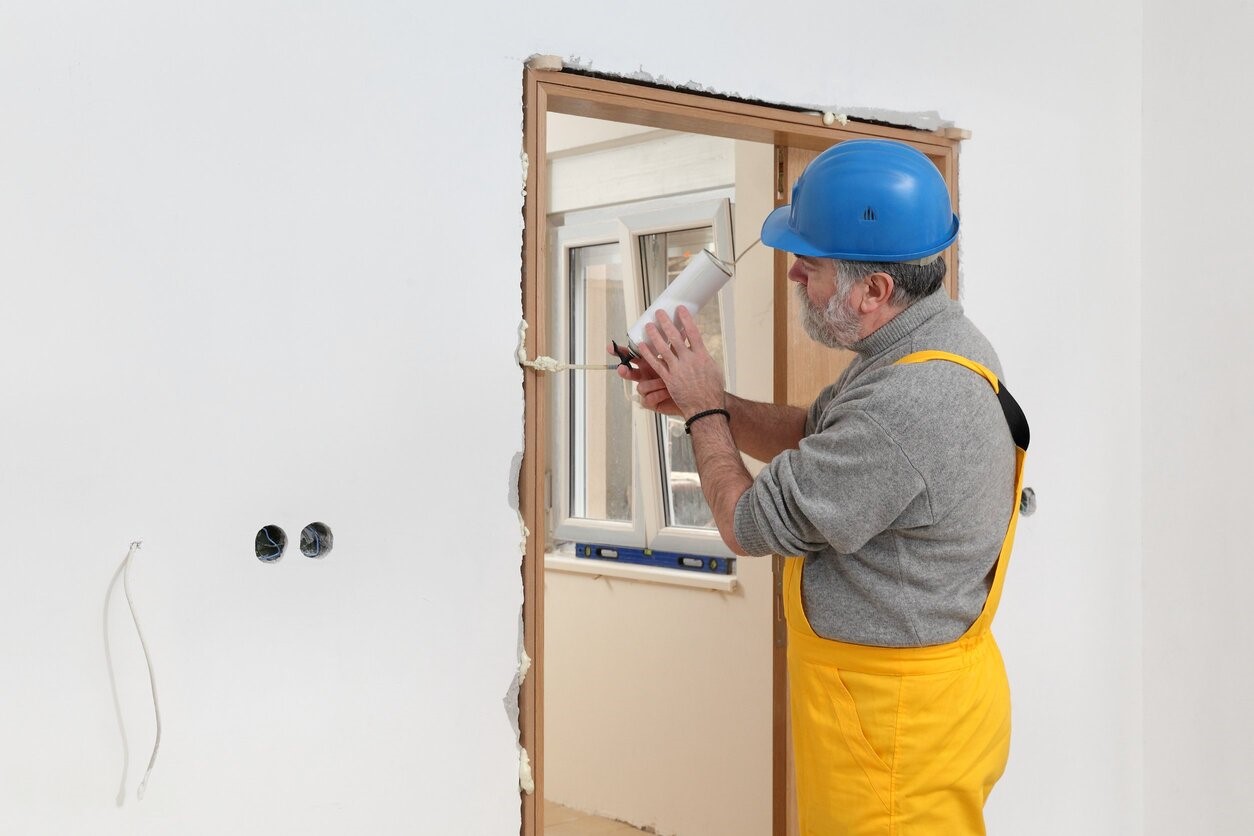 starszy mężczyzna z brodą, w niebieskim kasku i żółtym kombinezonie montuje ościeżnicę drzwiową, za pomocą pianki niskoprężnej