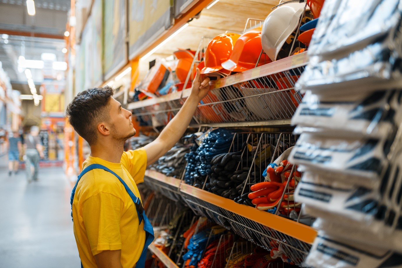 młody mężczyzna w żółtej koszulce i roboczym kombinezonie w sklepie budowlanym wybiera kask i robocze rękawice