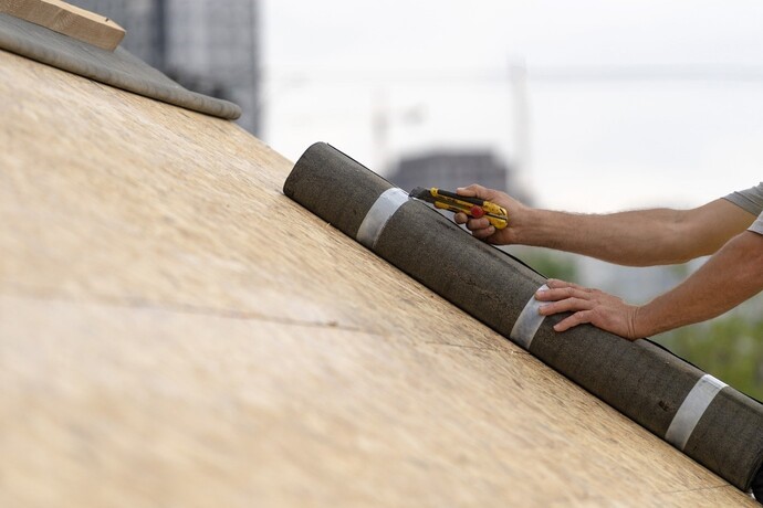 pracownik budowlany, na dachu nowoczesnej konstrukcji budynku, rozpakowuje rolkę z membraną dachową, za pomocą noża introligatorskiego