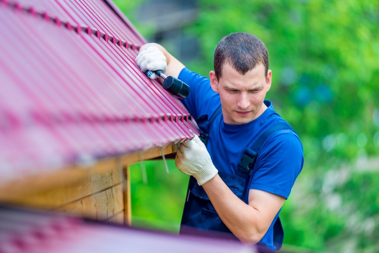 Młody mężczyzna w niebieskim, roboczym kombinezonie naprawia dach za pomocą narzędzi dekarskich